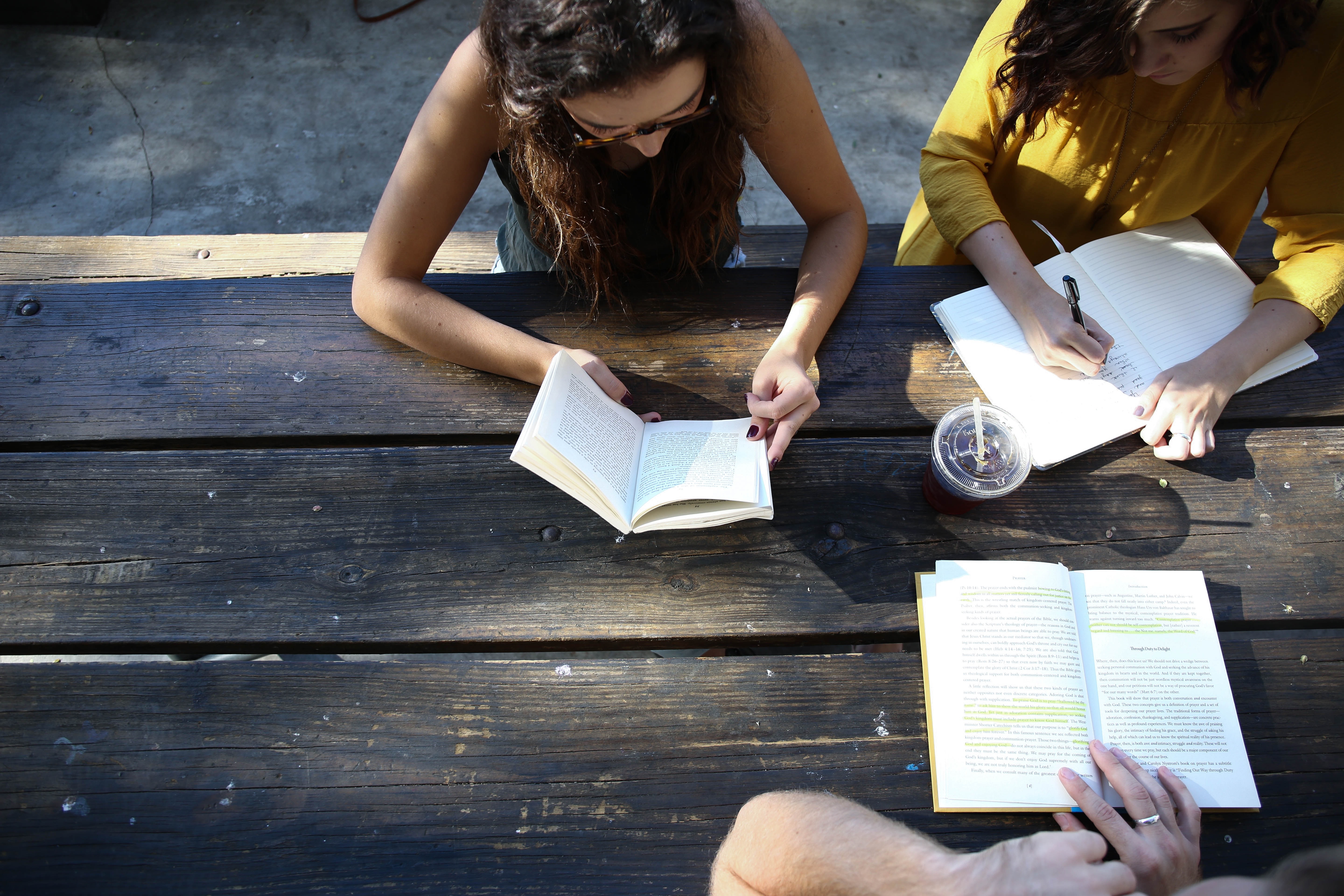 And overhead shot perspective with three people sitting at an outdoor old wooden park bench. On the top of the image are two young women about university age. The young woman on the left appears to be reading from a small book and the young woman on the right appears to be taking notes in a larger note book. There is a clear large plastic cup with lid and straw in front of the young women on the right. There is an older male person sitting across at the bottom of the screen has larger book open with some yellow highlighted lines 